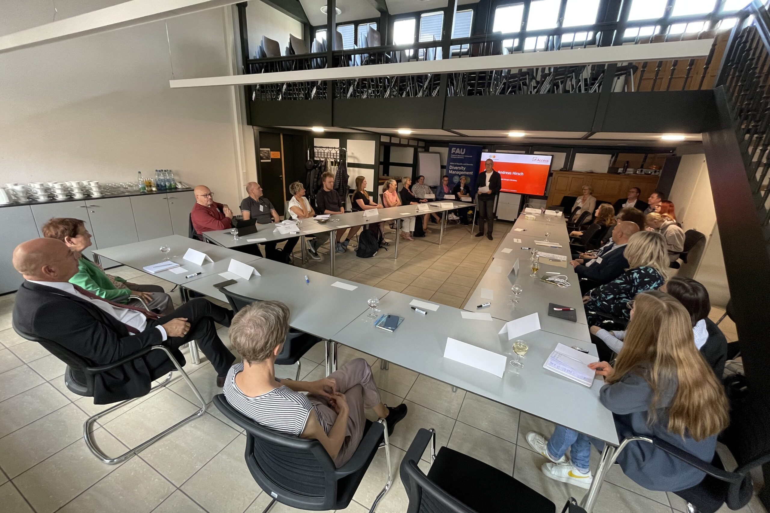Mehrere Personen sitzen an einem U-förmigen Konferenztisch. Eine Person hält einen Vortrag.