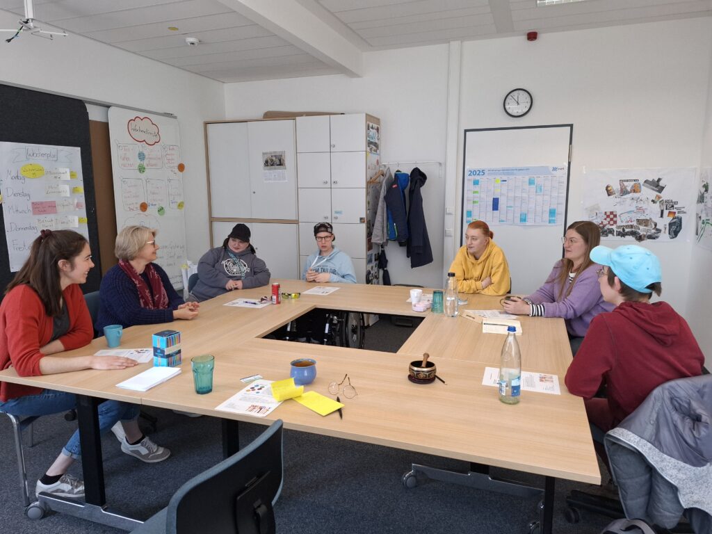 Auf dem Foto sitzen Qualifizierungstrainerin Lisa Plank, Projektleitung Claudia Drechsel und die vier angehenden Bildungsfachkräfte Ines Wolf, Marvin Voit, Peter Kutzi und Nadine Zitzmann gemeinsam mit Harriet Ziegler (Diversity Management der FAU) am Tisch im Qualifizierungsraum und unterhalten sich.