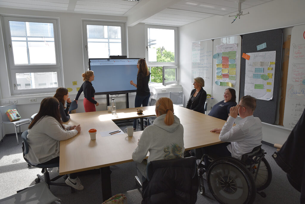 Zu sehen ist eine Alltagsszene im Qualifizierungsraum. Die Qualifizierungstrainerinnen Lisa Plank und Sina Zauner stehen am Smartboard. Weitere Teammitglieder sitzen an einem Tisch und wenden sich den Qualifizierungstrainerinnen zu.