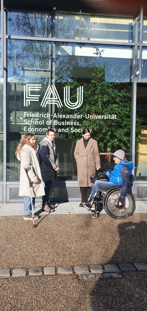 Qualifizierungstrainerin Sina Zauner, angehende Bildungsfachkraft Nadine Zitzmann, studentische Hilfskraft Donata Gaupp und angehende Bildungsfachkraft Marvin Voit befinden sich vor einem Gebäude der Friedrich-Alexander-Universität. 