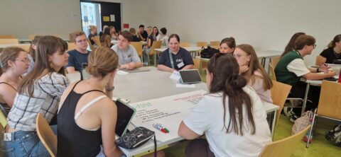 Eine Gruppe von Menschen sitzt um einen Tisch. Sie sind über Materialien gebeugt und scheinen an etwas zu arbeiten.