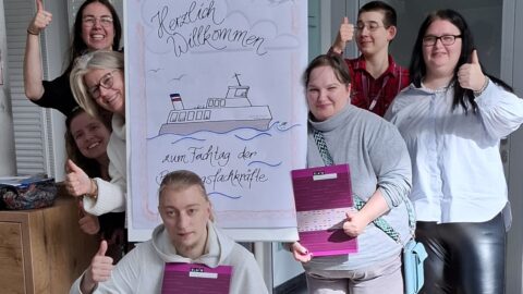 Eine Gruppe von sieben Personen steht um ein Flipchart. Auf dem Flipchart steht Herzliche willkommen zum Fachtag der Bildungsfachkräfte. Sie Personen um das Chart recken den Daumen nach oben und lachen.