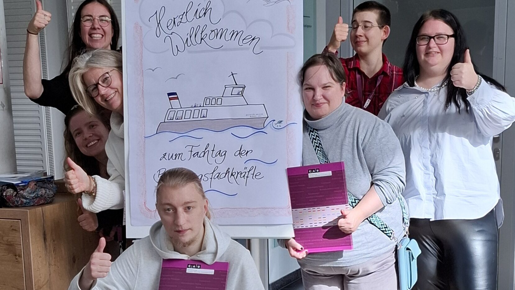 Eine Gruppe von sieben Personen steht um ein Flipchart. Auf dem Flipchart steht Herzliche willkommen zum Fachtag der Bildungsfachkräfte. Sie Personen um das Chart recken den Daumen nach oben und lachen.