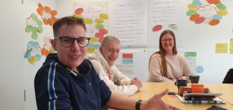 Drei Personen sitzen um einen Tisch. Sie wirken entspannt. Im Hintergrund ist ein Whiteboard mit vielen bunten Zetteln zu erkennen.