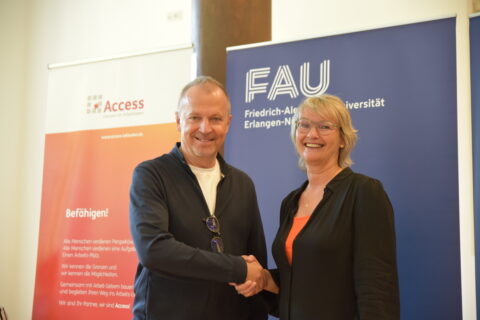 Ein Mann und eine Frau schütteln sich die Hand. Im Hintergrund sind auf Bannern die Logos der Friedrich-Alexander-Universtität Erlangen-Nürnberg und von Access - Inklusion im Arbeitsleben zu erkennen.